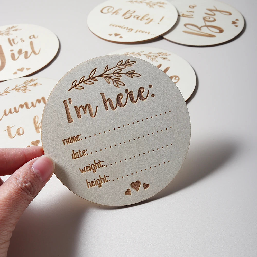 A hand holding a round wooden birth announcement with I'm here and spaces for name, date, weight, and height.