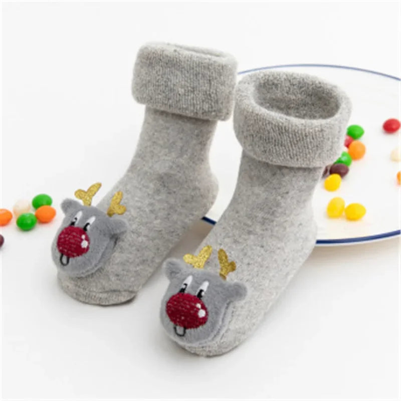 A Pair Of Gray Christmas Baby Socks With Reindeer Design With Plates Of Candies In The Background