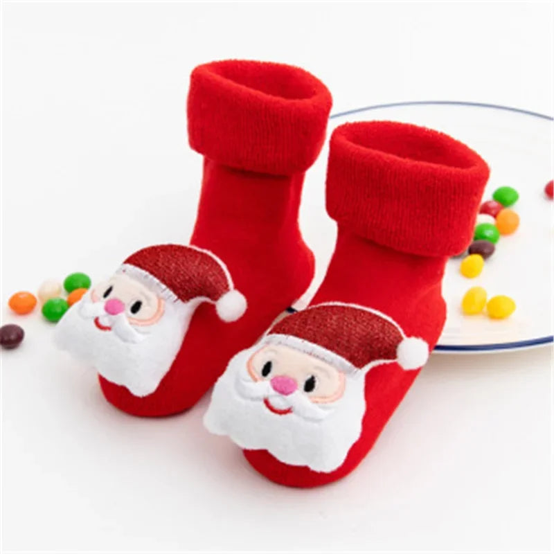 A Pair Of Red Christmas Baby Socks With Santa Clause Design With Plates Of Candies In The Background